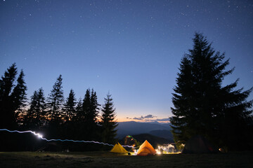 Bright illuminated tourist tents glowing on camping site in dark mountains under night sky with sparkling stars. Active lifestyle concept