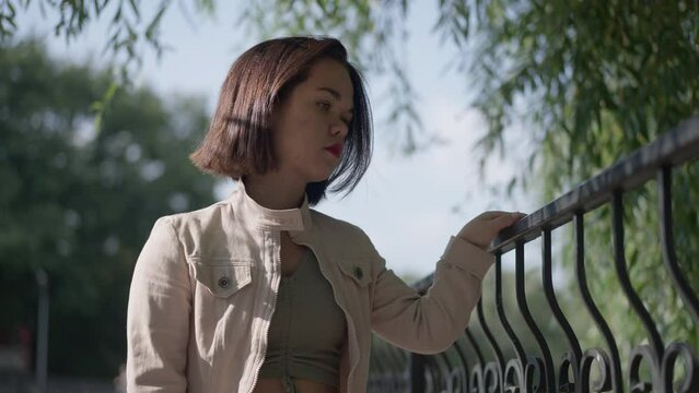 Thoughtful sad woman with dwarfism strolling on bridge in spring summer park looking away. Portrait of frustrated depressed Caucasian little person thinking leaving in slow motion