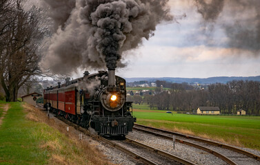 Fototapeta na wymiar An Antique Steam Passenger Train Traveling Thru Farmlands Puffing Lots of Smoke on a Cloudy Winter Day