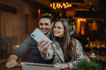 Two people young caucasian couple man and woman female and male friends or boyfriend and girlfriend taking selfie photos or making a video call while sitting at cafe copy space fun and romance