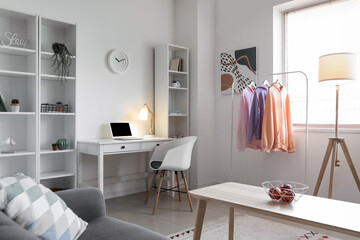 Interior of light room with modern workplace, rack and hoodies