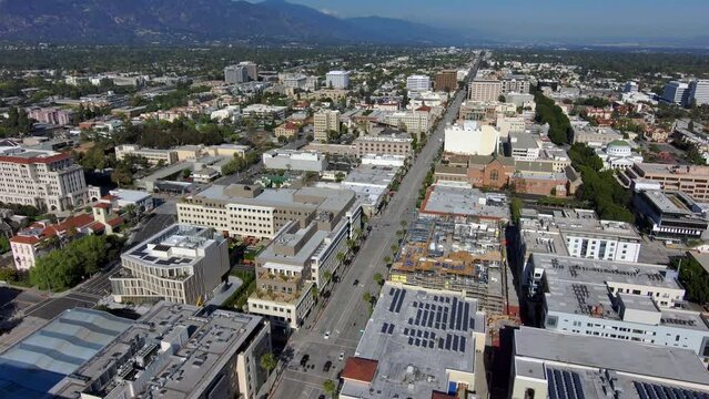 aerial footage of a long street with cars and trucks driving along the road surrounded by office buildings and shops with mountain ranges and lush green trees in Pasadena California USA