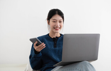 portrait of a happy casual asian woman holding laptop computer and hold smartphone  while sitting on a chair over white background