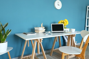 Workplace with modern laptop, books and vase with flowers near blue wall in room interior