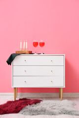 Glasses of wine and candles on chest near pink wall