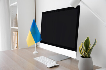 Modern workplace with computer, flag of Ukraine, houseplant and lamp near light wall