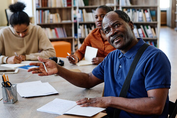 Portrait of mature Black man sitting at table in library asking teacher question about difficult...