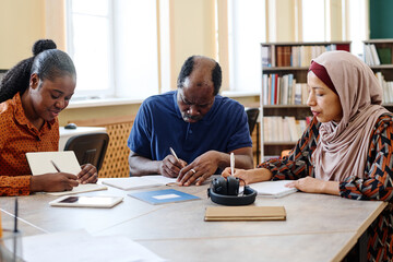 Group of three multi-ethnic immigrant students sitting at table in library doing writing task...