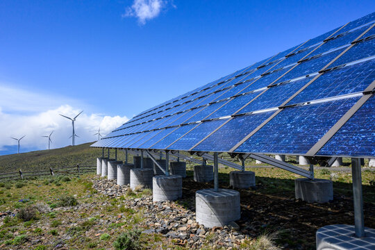 Commercial utility installation of blue photovoltaic panels on a sunny day, dry landscape with wind turbines in background, green power generation
