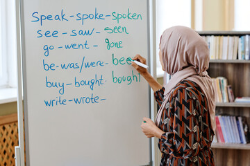 Unrecognizable woman wearing hijab standing at whiteboard doing task with irregular verbs during...