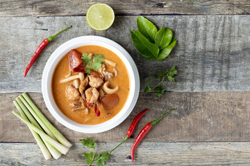 Soup Tom Yam with seafood, galangal, chili pepper, kaffir leaves, lime and cilantro in a white plate, on a wooden background. Top view.