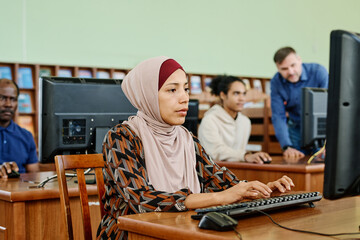 Portrait of modern Muslim woman wearing hijab sitting in university library working on desktop...