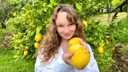 a fair-haired girl juggles lemons against the backdrop of a lemon tree in a tropical country, she is dressed in a white shirt she laughs and has fun High quality photo