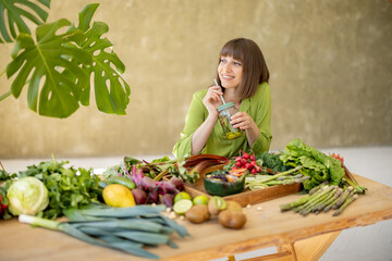 Young woman drinks from a bottle while sitting by the table full of fresh vegetables, fruits and...
