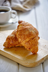 A crispy croissant with almond filling and hot black coffee on a wooden board on a light concrete table.