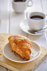 A crispy croissant with almond filling and hot black coffee on a wooden board on a light concrete table.