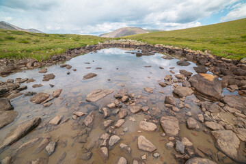 Scenic alpine landscape with small mountain lake on green grassy hill under cloudy sky. Dramatic mountain view to small lake with clouds reflection. Transparent water in alpine lake with stony bottom.