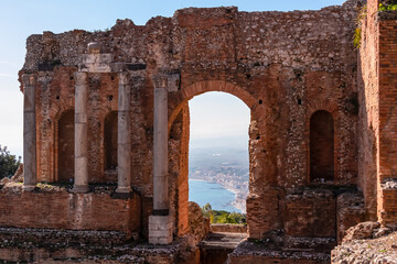 Panoramic view on the ruins of ancient Greek theater Taormina, island Sicily, Italy, Europe, EU. Looking through the red brick wall gate of the archeological ruins on the Mediterranean coastline - obrazy, fototapety, plakaty