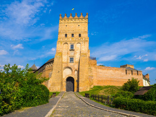 Fototapeta na wymiar Lutsk High Castle, also known as Lubart's Castle. Ukraine. Brick Tower and Walls. A part of defence wall of Lutsk Castle. The Entrance tower against the blue sky and with greenery under a brick wall.