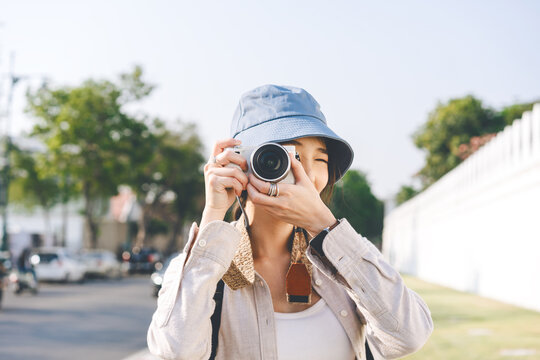 Attractive happy smile young adult asian woman traveller wear blue hat and backpack. Using camera for photo trip. People traveling in city lifestyle on day. Staycation summer trip concept.