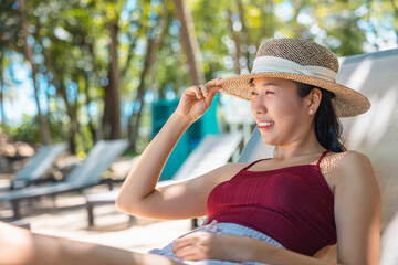 Model Asian woman wearing red swimsuit and straw hat lying on the sun lounger in sun by the sea. Beautiful girl relaxing at infinity luxury resort. Spa, wellness