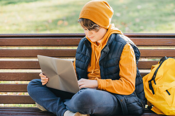 Child caucasian teen boy in yellow beany hat and backpack sitting on a bench outdoors using laptop....