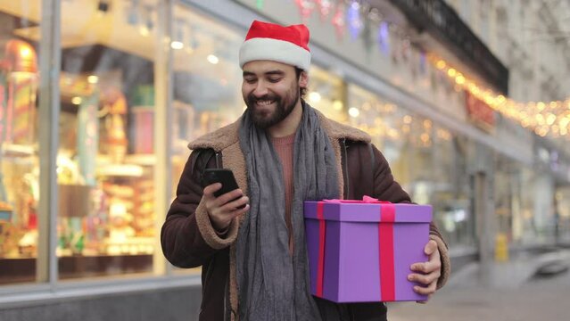 Happy young man in santa hat using modern smartphone for taking picture of christmas gift box. Caucasian bearded guy standing on street and chatting online on mobile.