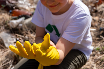 Young blonde white caucasian boy with a recycling symbol on his t-shirt putting on yellow gloves on the foreground of plastic in the environment