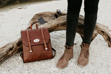 Autumn collection for hiking. Leather backpack, brown boots, sunglasses and a phone. Outdoor fashion idea. Nature lover style by a lake. A woman's leg with boots. 