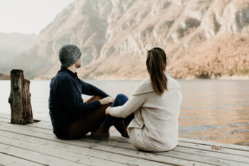 Fototapeta premium A couple in love is sitting on a wooden pier by a lake in Europe, in the Alps. They sit next to each other, facing the distance. Romantic sunset date scene. Travel destination idea, relaxing scenery.