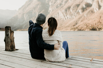 A couple is sitting and cuddling on a wooden pier at lake Bohinj. The man is pointing on something in the distance. Autumn travel destination. Active lifestyle, hiking outdoor, resting by the water.