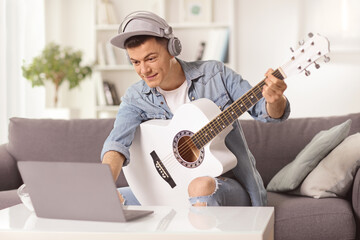 Guy sitting on a sofa at home playing an acoustic guitar and looking at a laptop computer