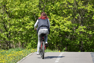 Girl in jeans with red dyed hair riding on a bicycle in a green park. Woman cyclist, spring or summer leisure