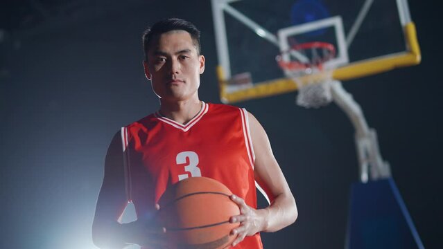 Portrait of an asian man basketball player, holding a ball in her hands and serious looking at the camera, basketball championship, 4k slow motion.