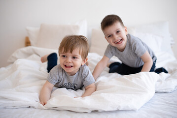 Smiling two boys in gray t-shirts lie and playing on white blanket on bed. Cheerful and happy childhood in family.  Exercise and play good for health. Together joyful and interesting. 