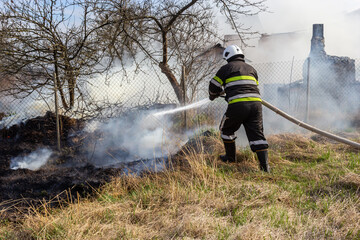 spring fire, burning dry grass near buildings in the countryside. Firefighter extinguishes the...