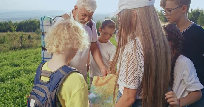 Side view of kids standing around, listening, looking, old man with rucksack holding map, talking. Tourists traveling, hiking together. Concept of tourism and team spirit.