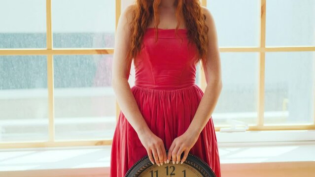 Sad cute barefoot gentle fantasy woman stand by window. Girl holds round wall clock hands, black number, white dial. Fairytale keeper time, red vintage elegant midi dress. Redhaired long curls on face