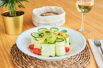 High angle of fresh zucchini rolls served in bowl with avocado slices and halves of cherry tomatoes in restaurant
