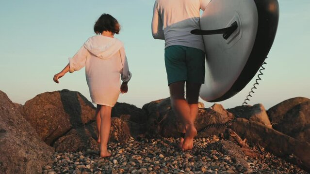 Family vacation by the sea. Father and daughter walk along the beach together. A man holds a supboard in his hands. Slow motion. Back view. The concept of surfing and sup boarding.