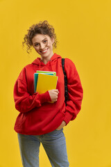 Front view of beautiful student in red khudi standing, looking at camera. Young blonde female with curly hair holding books, hand in pocket. Isolated on yellow studio background.