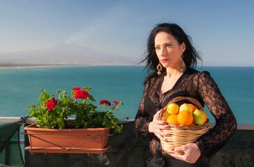 View of a typical mediterranean woman holding a wicker basket full of oranges and lemons in a panoramic balcony in Sicily, with blue sea and Mount Etna in the background - 506796769