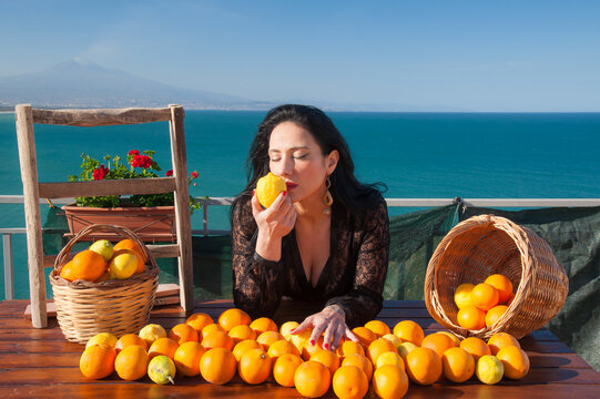 Mediterranean scenery: woman smelling oranges and lemons in a panoramic balcony in Sicily, with blue sea and Mount Etna in the background