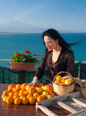 Mediterranean scenery: woman handling oranges and lemons in a panoramic balcony in Sicily, with blue sea and Mount Etna in the background