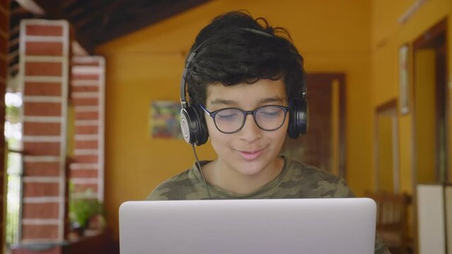 A young Indian Asian smiling male student wearing headphones and eyeglasses talking or interacting during online group class activity using a laptop. 
remote or distance education, technology concept
