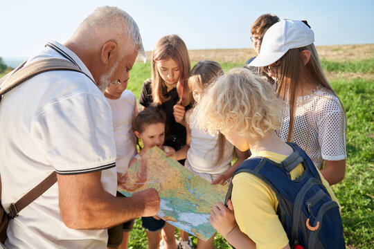 Close up of old man wearing white T-shirt and rucksack, showing map to children outside. Kids looking at map, searching route, camping in hills. Concept of mount hiking.