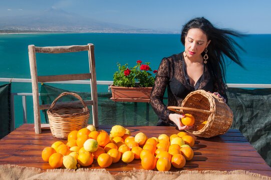 Mediterranean scenery: woman handling oranges and lemons in a panoramic balcony in Sicily, with blue sea and Mount Etna in the background