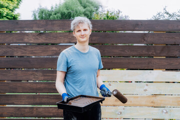 Portrait of young man worker paints with a roller a wooden board fence in the garden. DIY, Do it...