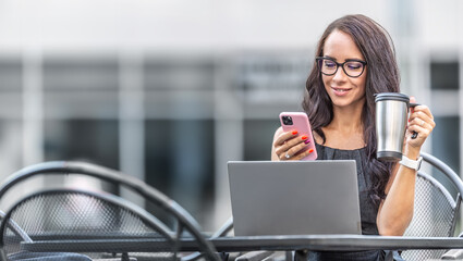 Good-looking woman drinks coffee from reusable mug and checks text messages on a phone with an open laptop in front of her