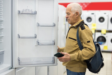 Naklejka na ściany i meble elderly grayhaired man pensioner looking refrigerator at counter in showroom of electrical appliance hypermarket department